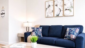 A living room with a dark blue sofa, two patterned pillows, a small white table with a plant and a book, and a wall-mounted lamp. - Stylish Suite • Downtown • Convention Center (Hot Springs)