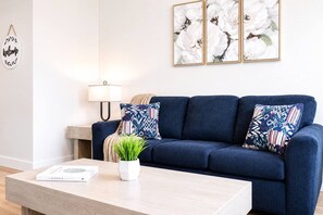 Een woonkamer met een donkerblauwe bank, twee gekleurde kussens, een klein wit tafeltje met een plant en een boek, en een wandlamp.