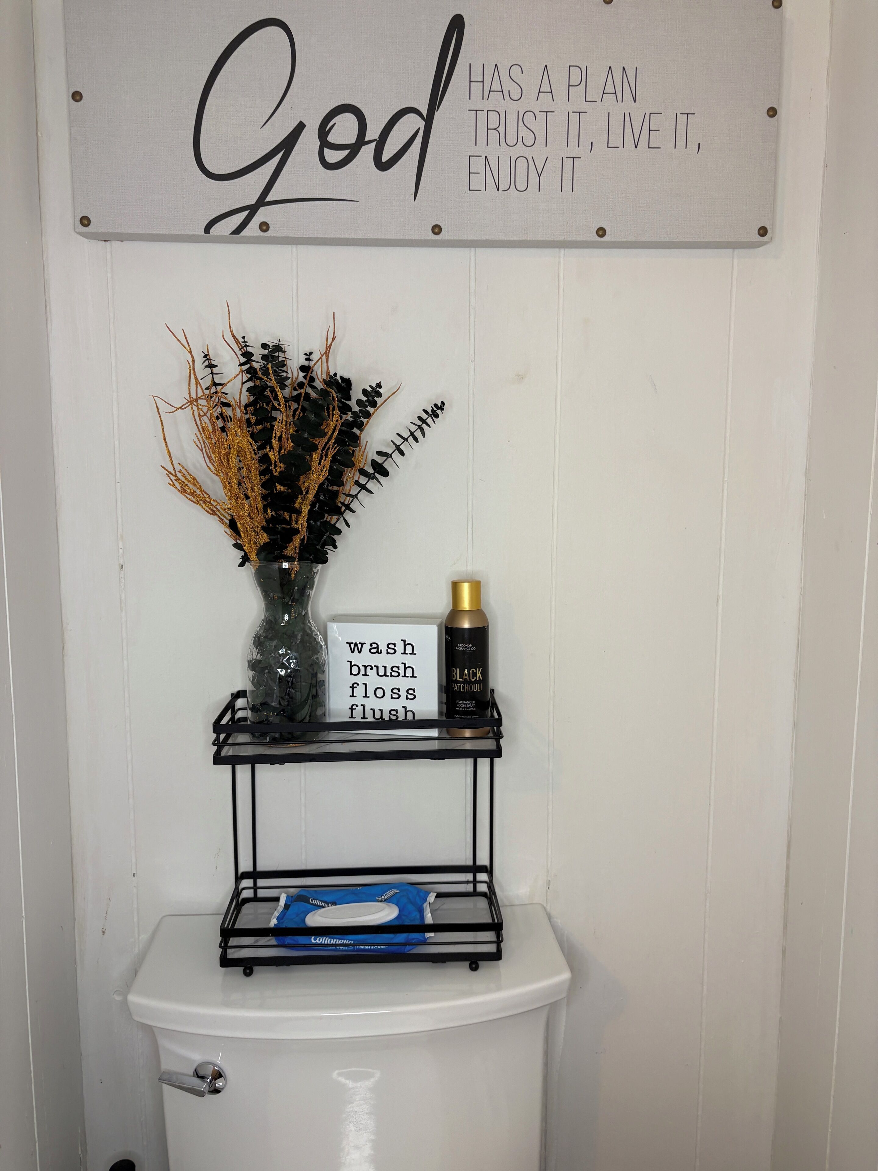 A bathroom with a toilet, a shelf with a sign, a vase with dried plants, and a bottle of Black Patchouli.