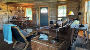L’intérieur d’un chalet rustique, avec des murs en bois, un coin repas, un espace salon confortable avec un canapé en cuir, et une petite table sur laquelle sont posés des livres et une lampe.