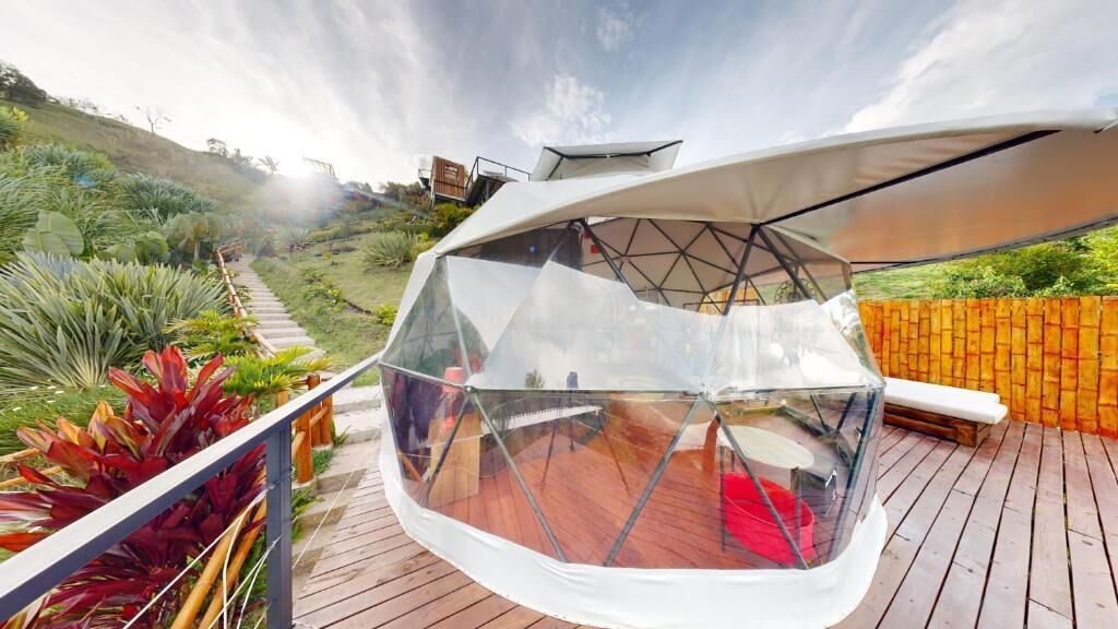 A geodesic dome structure with a transparent roof, surrounded by a wooden deck and lush greenery.