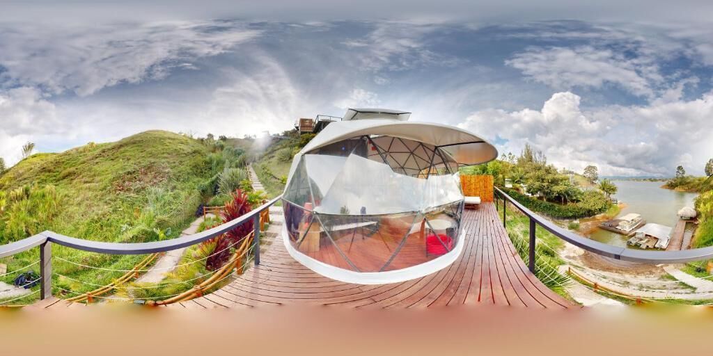 A dome-shaped structure with a transparent roof, surrounded by a wooden deck and a railing, overlooking a hilly landscape and a body of water.