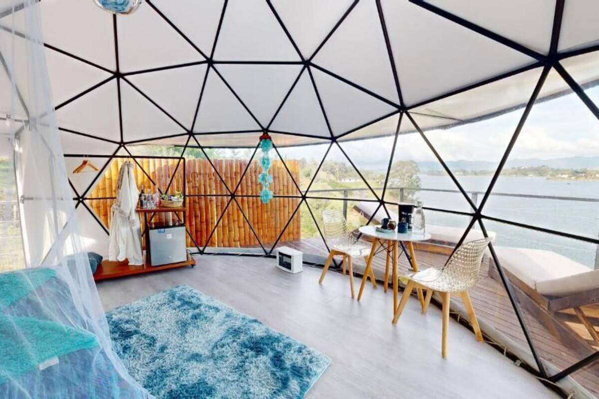 Una habitación en forma de cúpula geométrica con vista al agua, una terraza de madera y un área de estar con sillas y una mesa.