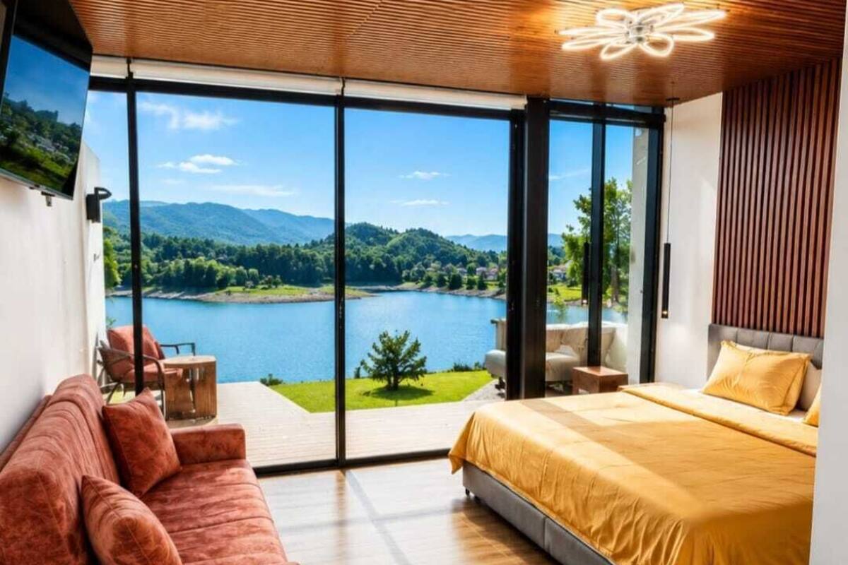 Un dormitorio con un ventanal grande, una cama, un sofá y vistas a un lago y montañas.