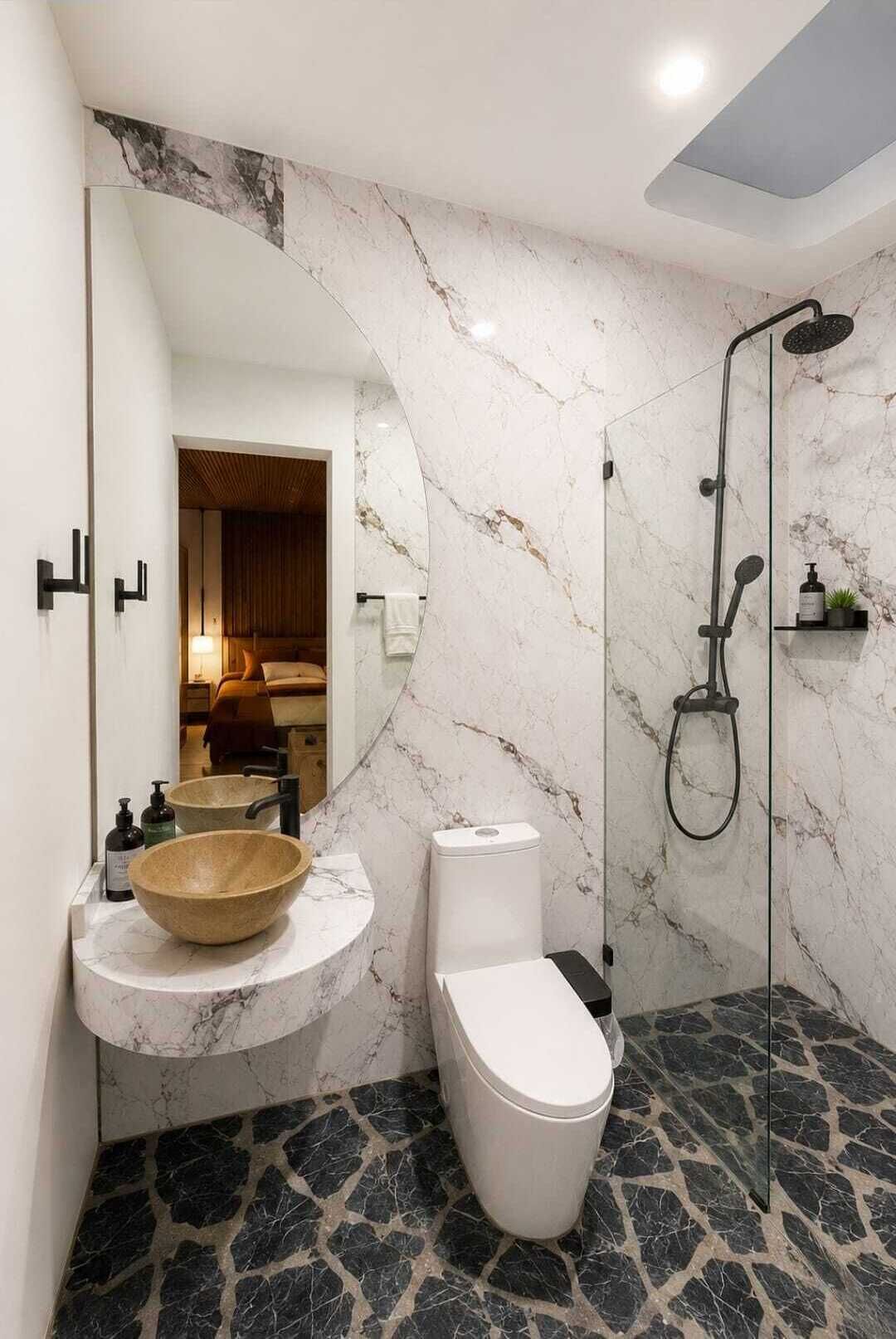 A bathroom with a marble vanity, a toilet, a shower, and a mirror reflecting a bedroom.