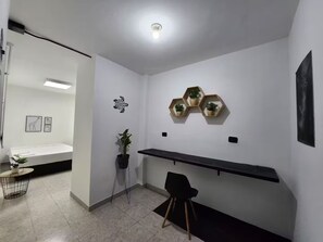 Ein modernes, minimalistisches Zimmer mit einem Bett, einem Schreibtisch und einem Sessel. An der Wand befinden sich Regale mit Pflanzen und ein Kunstwerk mit einer Schildkröte.