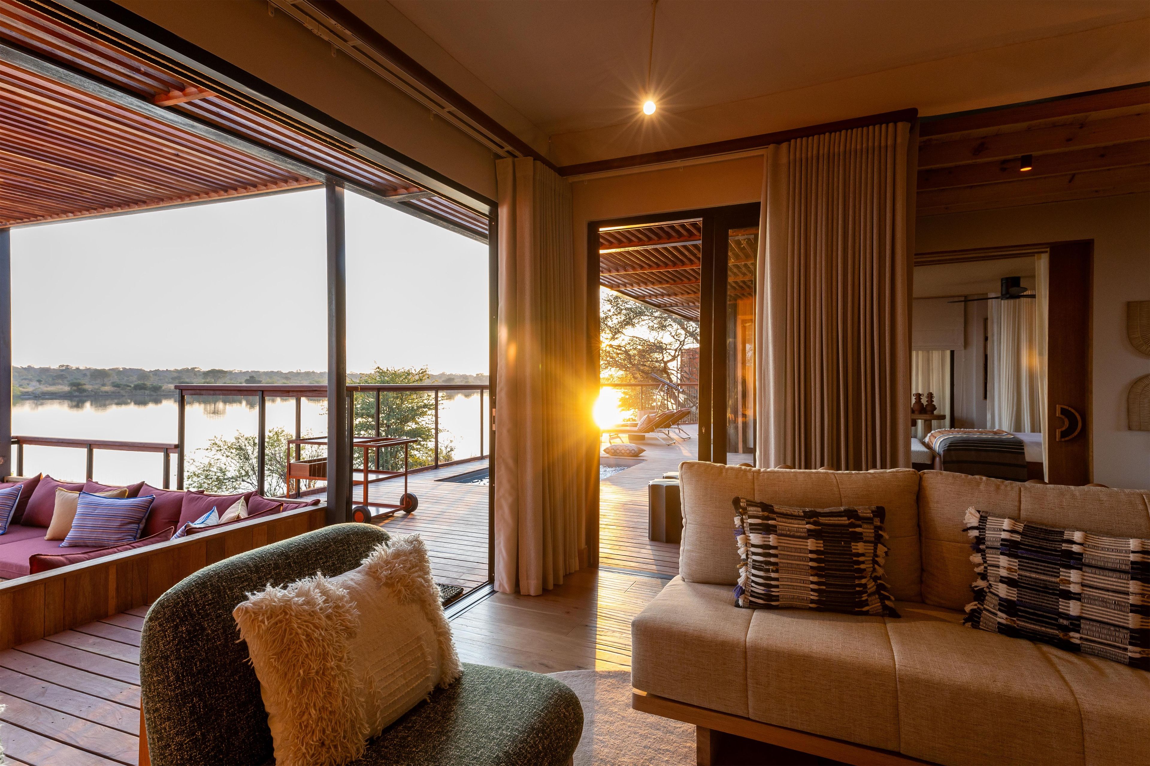 A cozy room with a large window overlooking a body of water, a wooden deck, and a seating area with cushions.