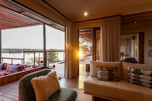 A cozy room with a large window overlooking a body of water, a wooden deck, and a seating area with cushions. - Anantara Tented Camp Kafue River (Kafue National Park)