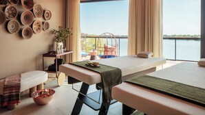 A spa room with two massage tables, a wall of decorative bowls, a small table with a plant, and a view of a body of water through large windows. - Anantara Tented Camp Kafue River (Kafue National Park)