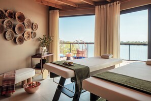 A spa room with two massage tables, a wall of decorative bowls, a small table with a plant, and a view of a body of water through large windows. - Anantara Tented Camp Kafue River (Kafue National Park)