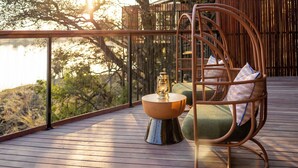 Eine Holzterrasse mit einem Korbstuhl, einem kleinen Tisch mit einer Laterne und Blick auf Bäume und ein Gebäude.