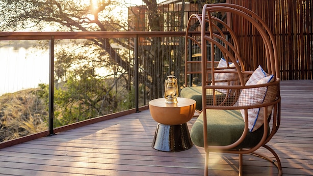 A wooden deck with a wicker chair, a small table with a lantern, and a view of trees and a building. - Anantara Tented Camp Kafue River (Kafue National Park)