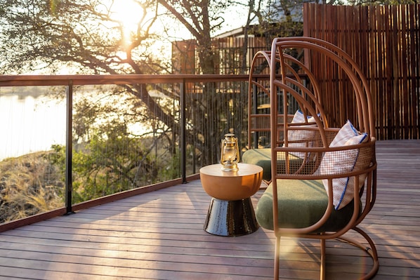 Una terraza de madera con una silla de mimbre, una mesita con un farolillo, y vistas a árboles y un edificio.