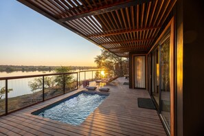A deck with a swimming pool, lounge chairs, and a view of a lake at sunset. - Anantara Tented Camp Kafue River (Kafue National Park)