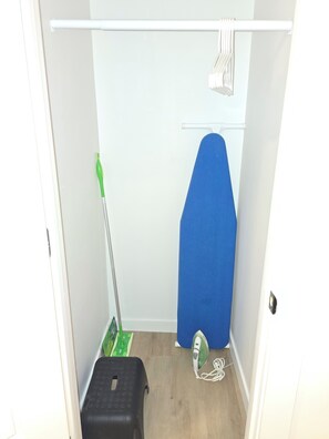 Ein kleiner, schmaler Raum mit einer weißen Tür, einem blauen Bügelbrett, einem grün-weißen Bügelbrett, einem schwarzen Koffer und einem Mop.