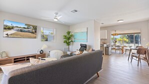 A modern living room with a grey sofa, a dining area with wooden chairs, a large window, and a ceiling fan. - Casa del Sol - Venice Island Beach Escape (Venice)