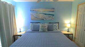 Une chambre à coucher avec un lit, un tableau représentant une scène de plage et une table de chevet avec une lampe.