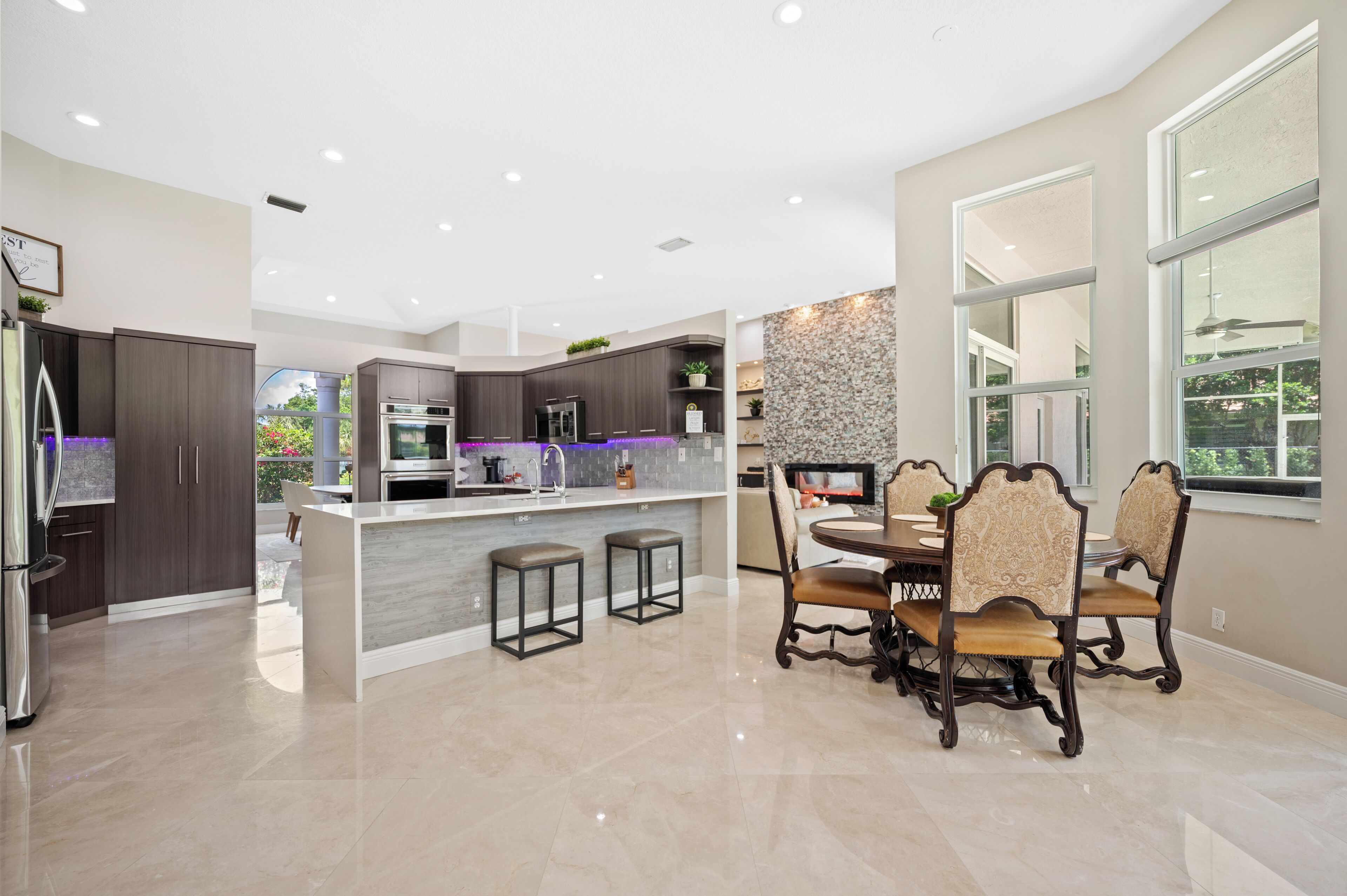 A modern kitchen with dark cabinetry, a central island, and a dining area with ornate chairs.