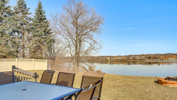 An outdoor patio with a table and chairs, a wooden deck, and a view of a lake with buildings in the background.