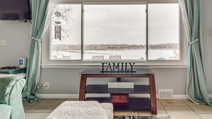 A living room with a large window, a television mounted on the wall, a small table with a sign, and a cozy chair.
