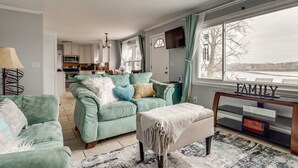 A living room with a teal sofa, a coffee table, and a TV. There is a kitchen in the background, a window with a view, and a ceiling fan.