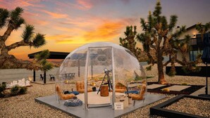 A transparent dome structure with seating inside, surrounded by desert vegetation and a sunset view. - Majestic Luxury Retreat | Pool ·Dome · Mini Golf (Joshua Tree)