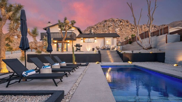A modern pool area with lounge chairs, a pool with blue lighting, and a house with a balcony in the background. - Majestic Luxury Retreat | Pool ·Dome · Mini Golf (Joshua Tree)