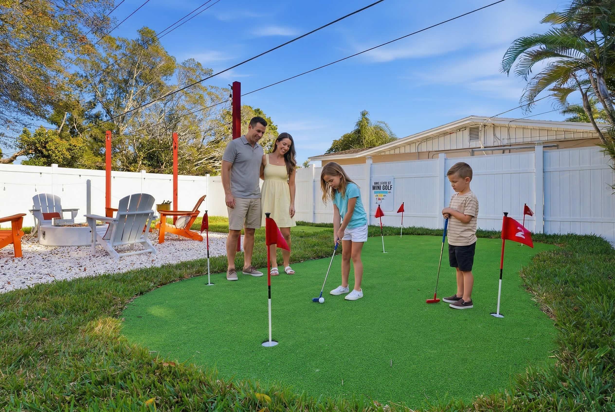 A family playing mini-golf outdoors with a white fence, chairs, and a building in the background.