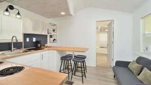 Een compacte keuken met een houten aanrecht, een spoelbak en een koffieapparaat. In de aangrenzende woonruimte staan vier barkrukken en een donkergrijze bank.