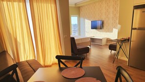 A modern living room with a sofa, a TV mounted on the wall, and a dining area with a table and chairs.