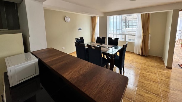 A modern kitchen with a built-in microwave, a wooden dining table with chairs, and a large window providing natural light. - Santa Ana Home, ideal para tu viaje en grupo o familia. (Cusco)