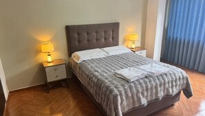 A neatly made bed with a grey blanket and white pillows, two bedside lamps, and a wooden nightstand with a drawer. - Santa Ana Home, ideal para tu viaje en grupo o familia. (Cusco)