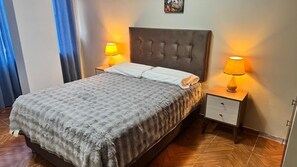 A neatly made bed with a grey patterned bedspread and white pillows, flanked by two bedside tables with lamps. The room has a wooden floor and a blue curtain on one side. - Santa Ana Home, ideal para tu viaje en grupo o familia. (Cusco)