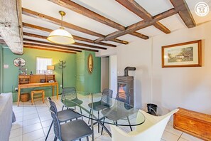 Un coin repas avec une table et des chaises en verre, un banc en bois, un poêle rustique et un mur vert agrémenté d’une cage à oiseaux et d’une étagère.