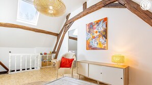 Une pièce avec des poutres apparentes, un radiateur blanc, un tableau abstrait aux couleurs vives, une chaise blanche avec un coussin rouge et une petite table avec une lampe.