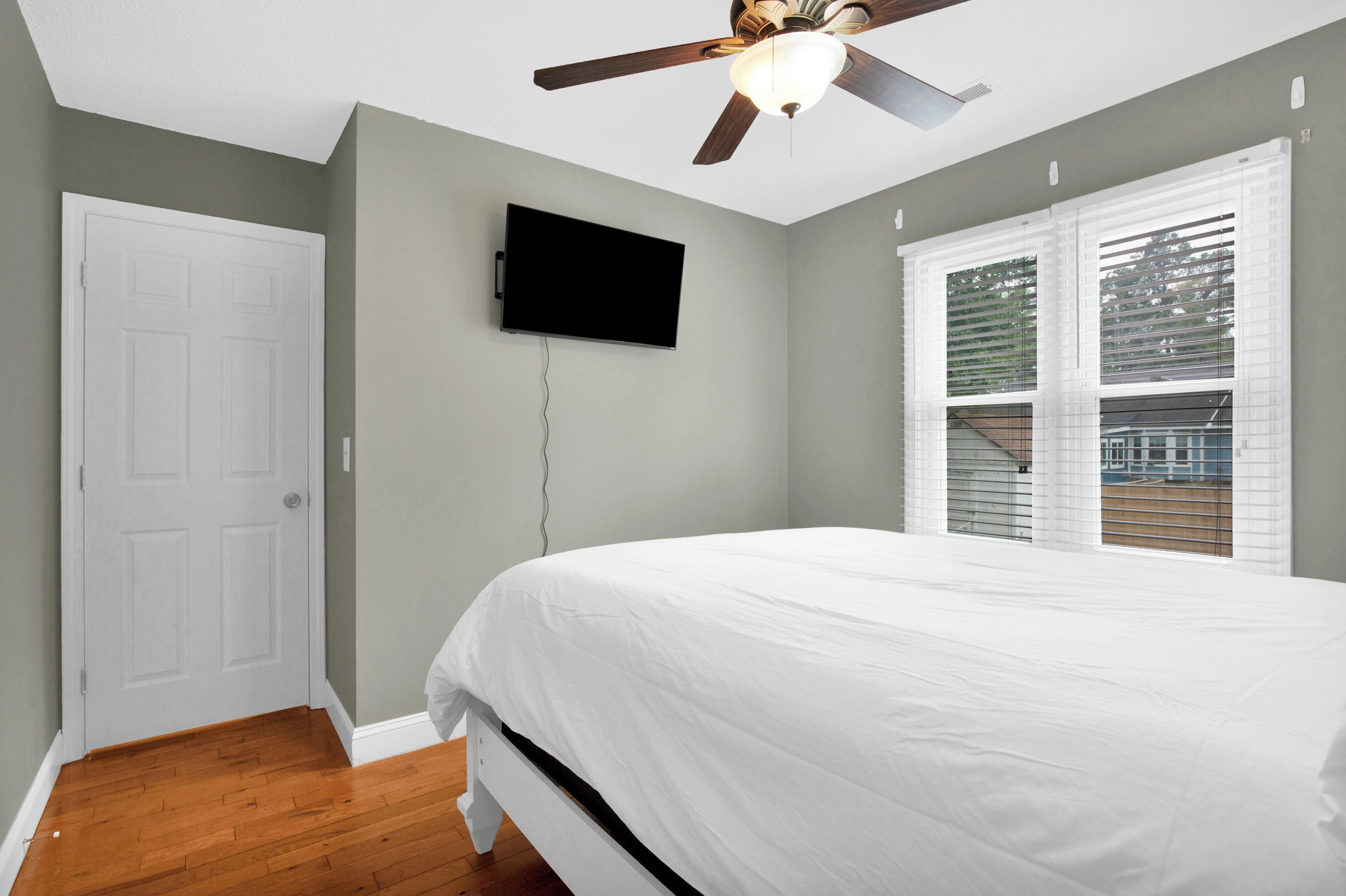 Un dormitorio con cama, ventilador de techo, televisor montado en la pared y ventana con persianas.
