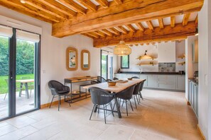 A modern kitchen with a wooden ceiling, a dining area with a table and chairs, and a view of a garden through large sliding doors.