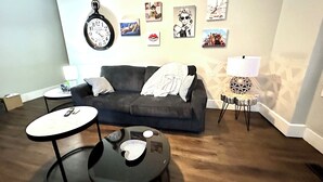 A living room with a dark-colored sofa, two round tables, a wall clock, and several framed pictures on the wall.