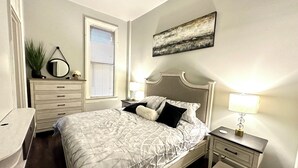 A neatly arranged bedroom with a bed, bedside tables, a dresser, and a window with blinds.
