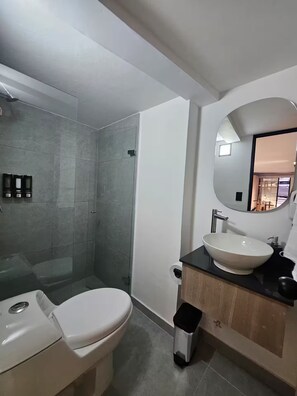 A modern bathroom with a glass-enclosed shower, a white toilet, and a vanity with a sink and mirror. - Naranjo Cozy Loft w/AC in Laureles (Medellín)