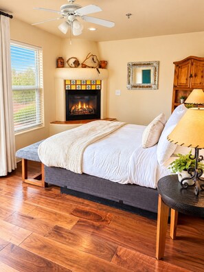 A bedroom with a bed, a fireplace, a ceiling fan, a window with blinds, and a wooden floor.