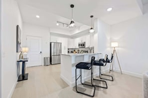 A modern kitchen with white cabinets, stainless steel appliances, and a central island with bar stools. - RIVER FLY-IN: Penthouse w/ Views & Amenities (Merritt Island)