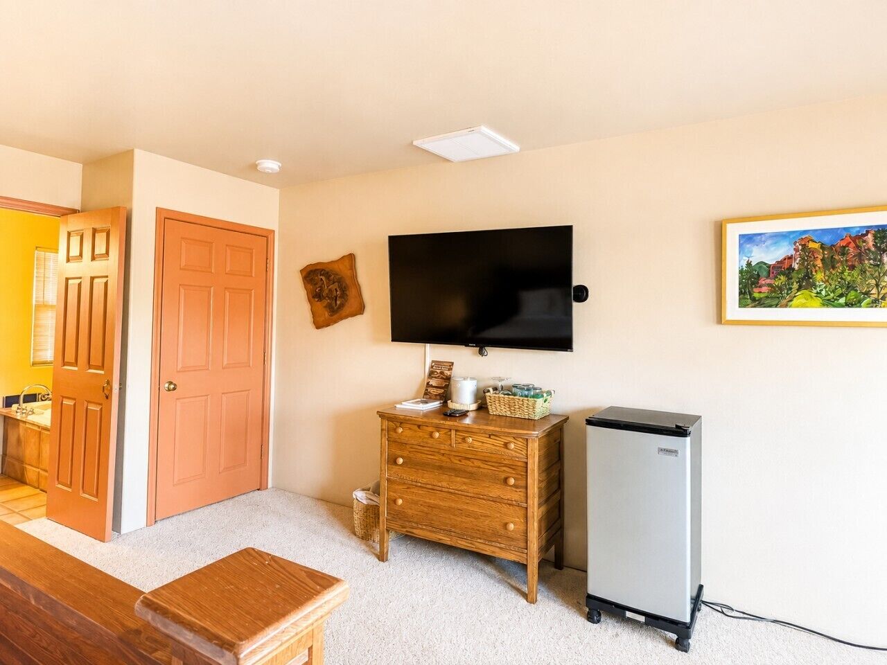 A room with a wooden dresser, a television mounted on the wall, and a refrigerator.