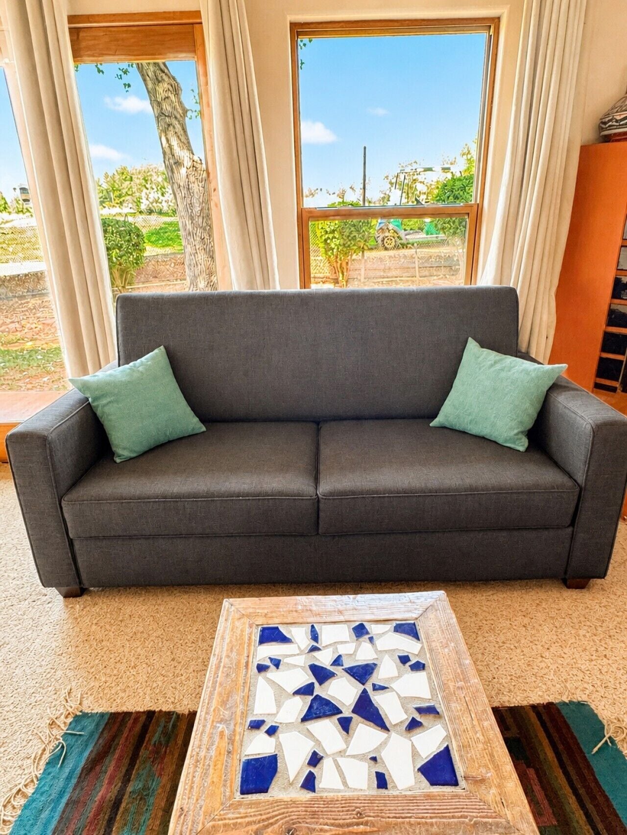 A dark grey sofa with two green cushions in a room with large windows, a wooden coffee table with a mosaic top, and a carpeted floor.