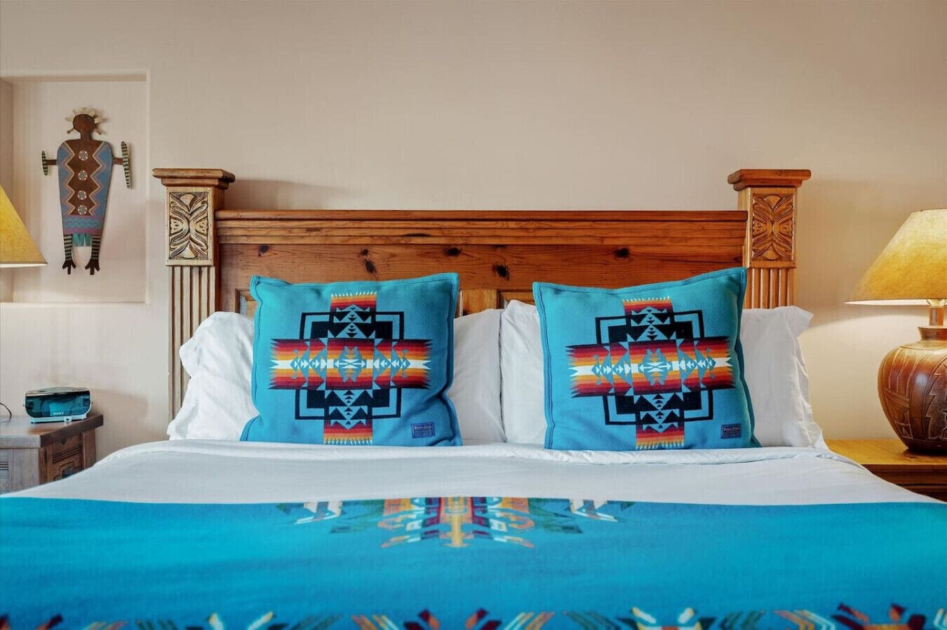 Una cama con dos almohadones decorativos, un cabecero de madera y una mesita de noche con una lámpara.