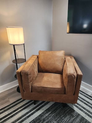 A brown leather armchair in a corner of a room with a striped rug, a standing lamp, and a flat-screen TV mounted on the wall.