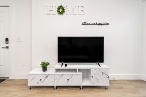 A modern living room with a flat-screen TV, a white entertainment unit with drawers, a small potted plant, and a 'HOME' sign with a wreath. - Stylish 1st-Floor Suite Near Convention Center (Hot Springs)