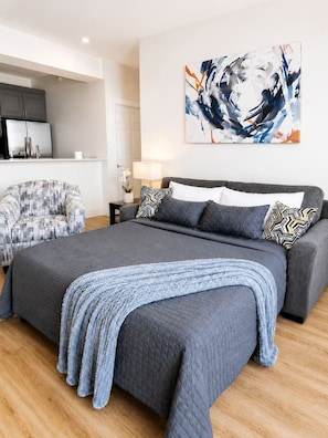 A modern bedroom with a large bed, a grey sofa, a small table with a lamp, and a wall-mounted abstract painting. - Stylish 1st-Floor Suite Near Convention Center (Hot Springs)