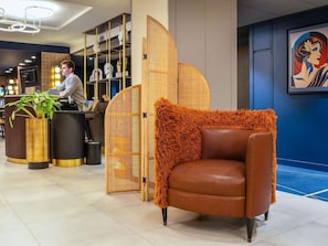 A lobby with a reception desk, a brown leather armchair, a large woven partition, and a framed artwork on the wall. - Ibis Styles Saint Quentin (Saint-Quentin)