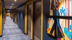 A hotel corridor with doors on the right, a patterned carpet, and a colorful mural on the wall. - Ibis Styles Saint Quentin (Saint-Quentin)